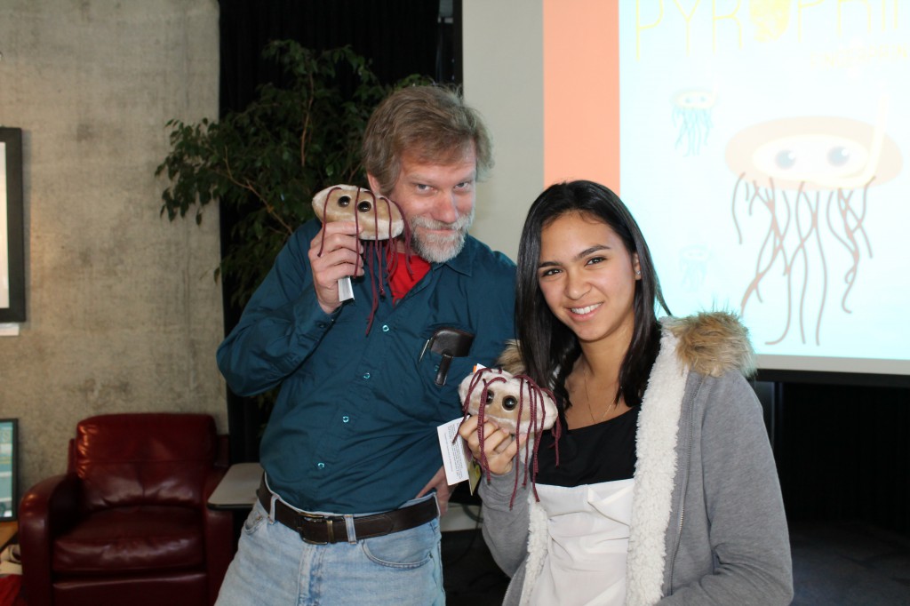 A bearded man and a young woman stand next to each other, smiling and holding small stuffed animal models of E. coli, which are fuzzy, oval-shaped and have two big eyes and little tentacles.