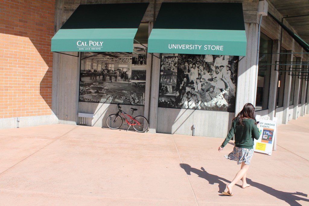 A photo of two windows, decorated with black and white photos of student groups. The green awnings above them say "Cal Poly" and "University Store." A women in a green sweater is walking by the photos.