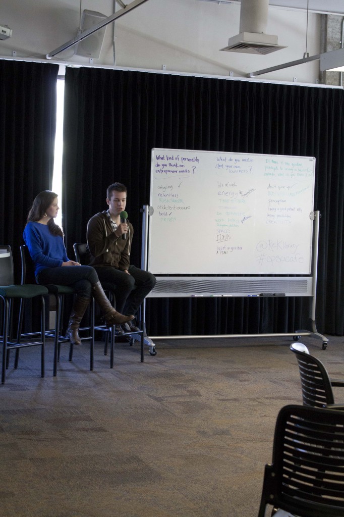 Jessie, a woman in a blue sweater, and Mark, in a brown sweater, sit on tall chairs next to a whiteboard where people wrote their assumptions about entrepreneurs.