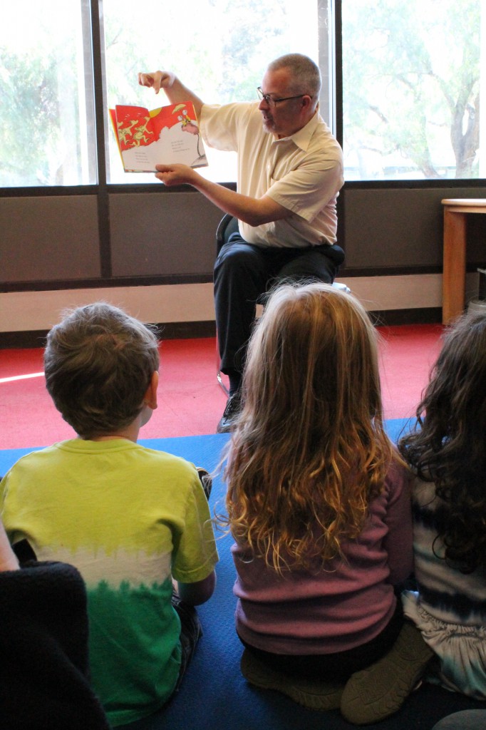 A man in glasses and a yellow shirt holds up One Fish, Two Fish, Red Fish, Blue Fish and points to the illustration. In front of him sit several young children.