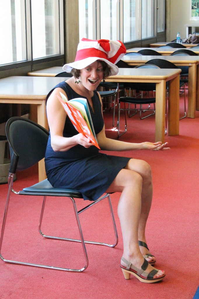 A woman in a blue dress and red and white striped Cat in the Hat hat sits on a chair reading Green Eggs and Ham with animated gestures.