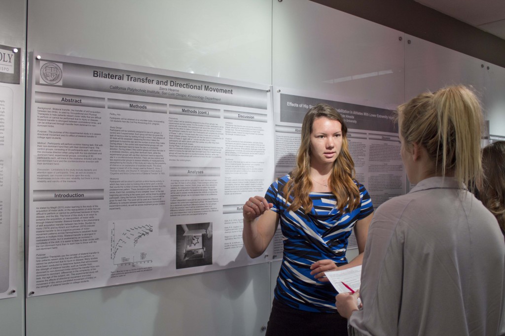 A woman with long hair and a blue, black and white top stands in front of a poster proposing a kinesiology research project. She is talking to a blonde woman standing in the foreground with her back to the camera.