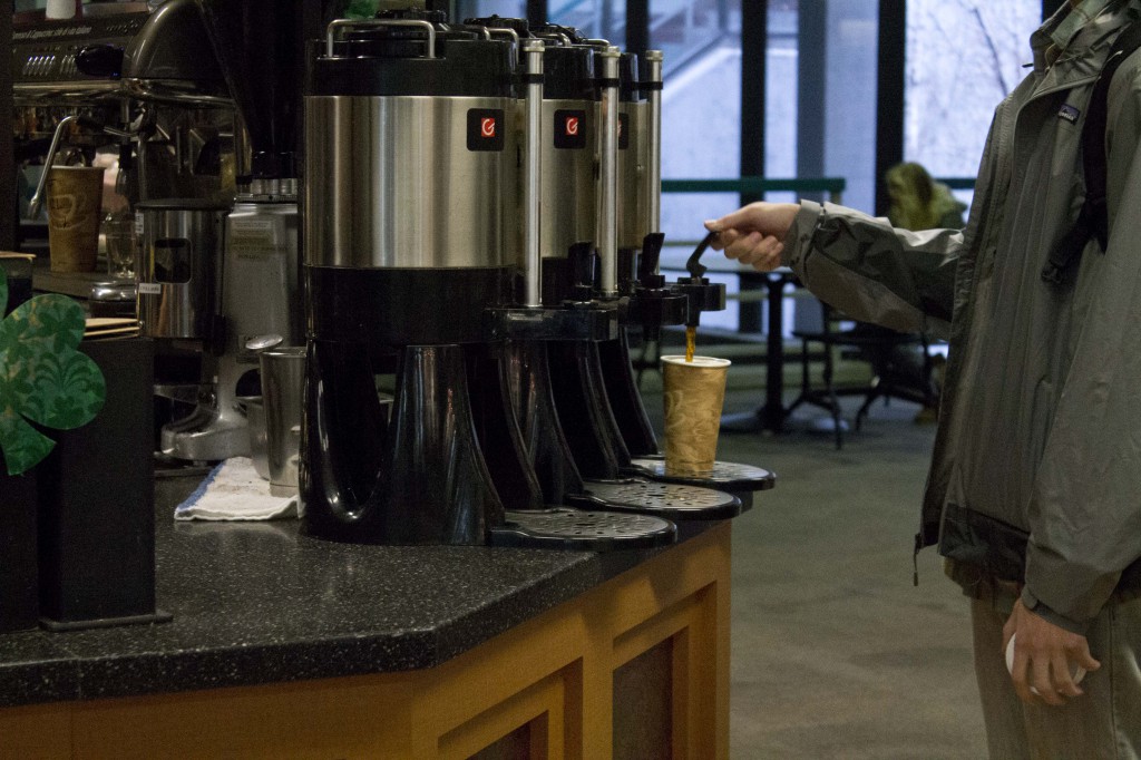 Three coffee pots sit on a counter; a man in a rain jacket fills a paper cup at the farthest coffee pot. We only see his arms and torso.