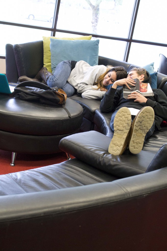 A man and woman nap next to each other on a half-circle leather couch. They are surrounded by backpacks, books, laptops and other school supplies.