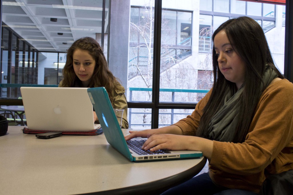 Two women sit at a table, working on laptops. They wear scarves and jackets. Behind them, we see out a window that it is gray and damp outside.