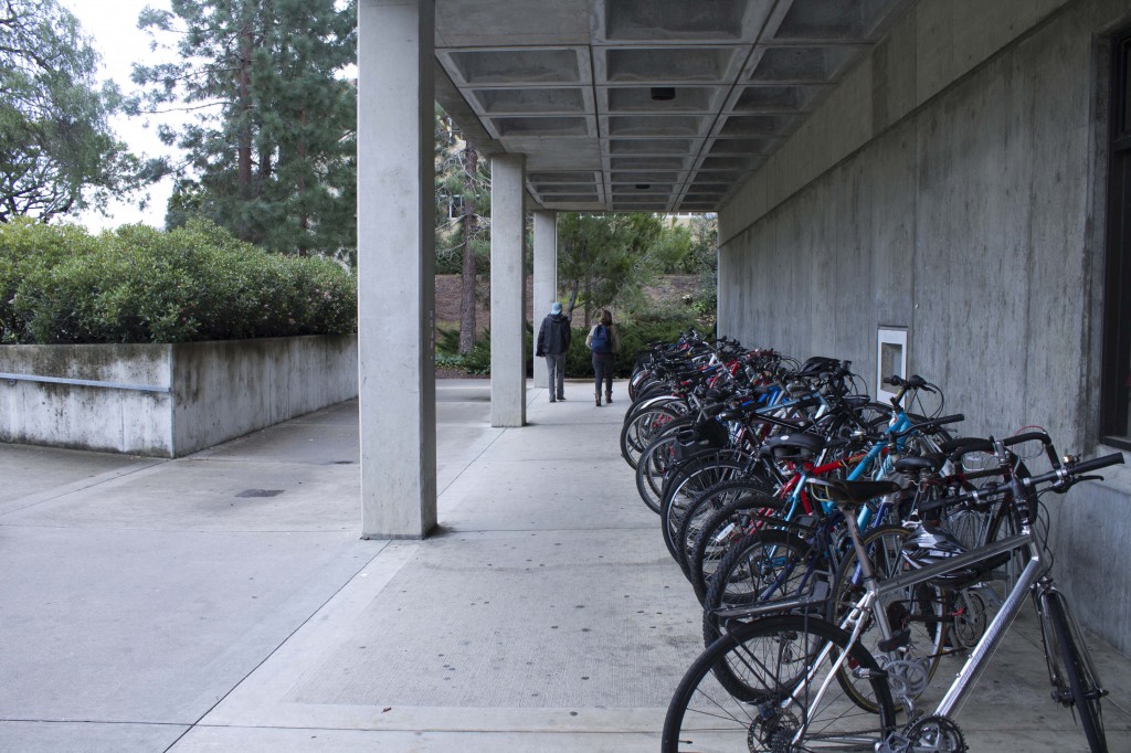 A row of bike racks, maybe 50 yards, crowded with bikes underneath a concrete overhang in front of the library.