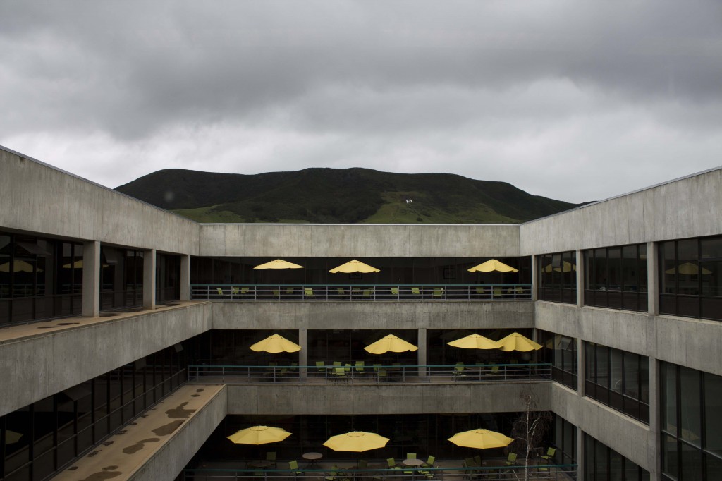 A photo from the 5th floor of Kennedy Library. We are looking out onto several stories of the cement and glass building. The balconies are decorated with tables with yellow umbrellas. In the background, there are gray rainclouds over a green mountain with a white letter P painted on it.