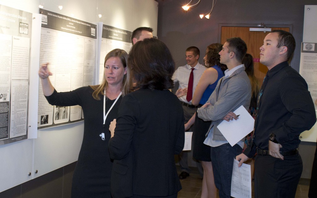 A blonde woman in black raises a hand to gesture as she explains her project to another woman. Behind them, at least six students in formalwear look at project posters on the wall.