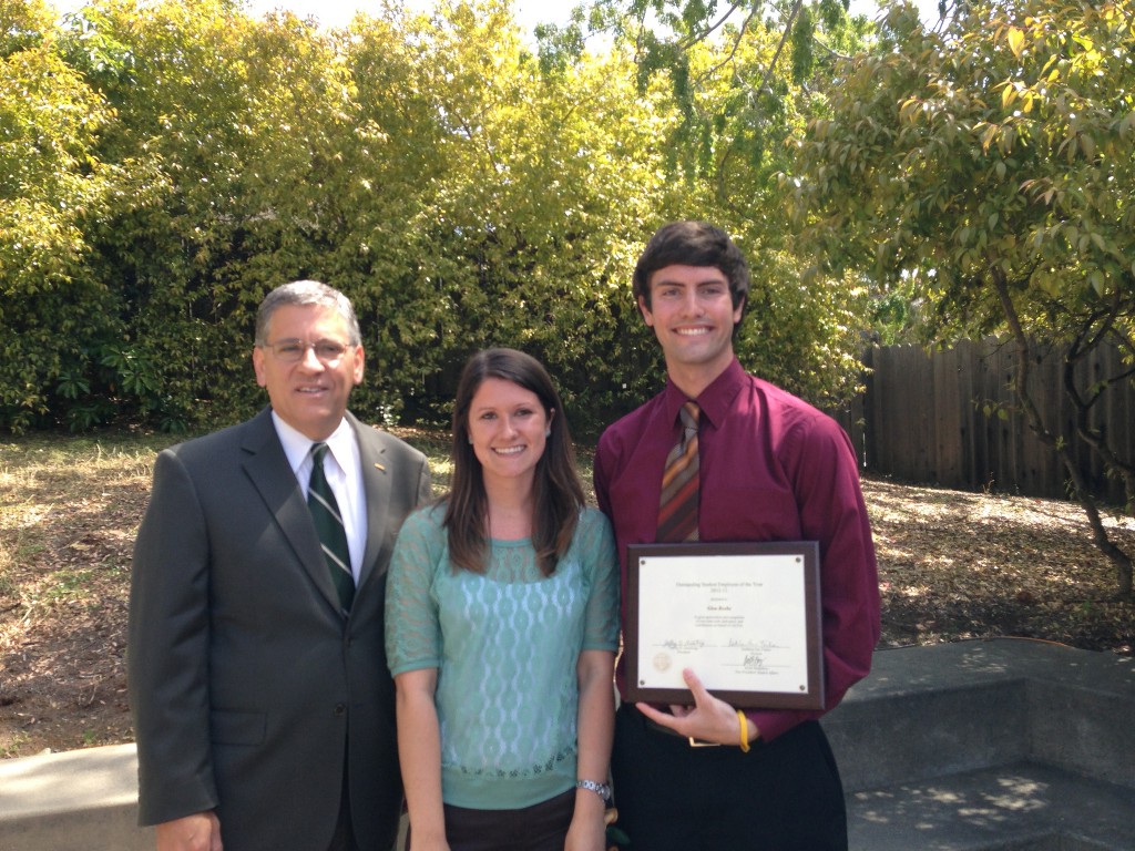 A man in a suit and glasses stands next to a young woman in turquoise and a young man in a burgundy button up and tie. The young man holds a plaque for Outstanding Student Employee of the Year. They are outside and it is sunny.