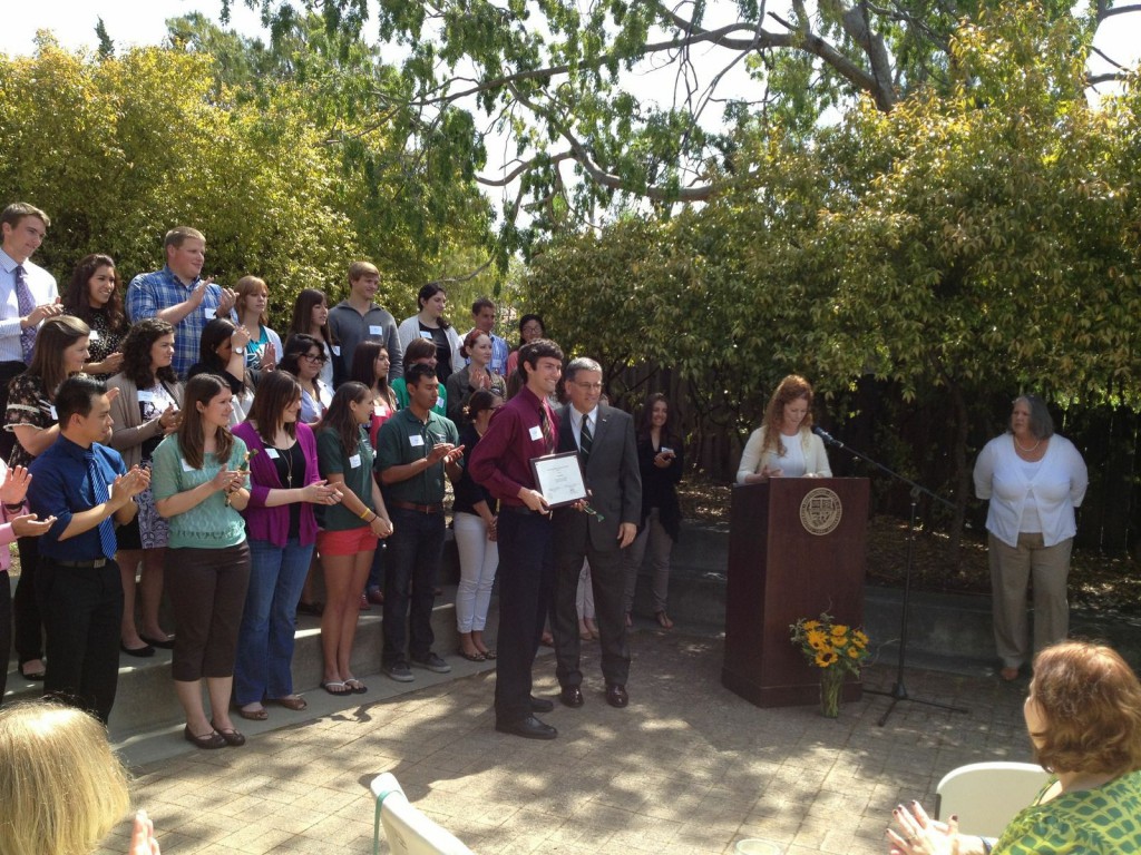 A young man in a tie stands next to the Cal Poly president in a suit, holding a plaque, while a crowd of student employees behind them applaud. A woman stands to their left at a podium reading.