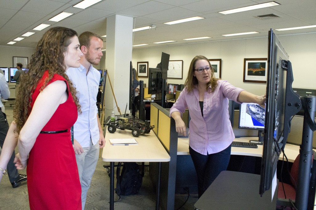 A woman in a red dress and a man in khaki stand facing a TV monitor. Another woman in a purple shirt stands in front of the monitor, pointing to the screen and explaining to them. The screen is at an angle so its contents aren't visible.