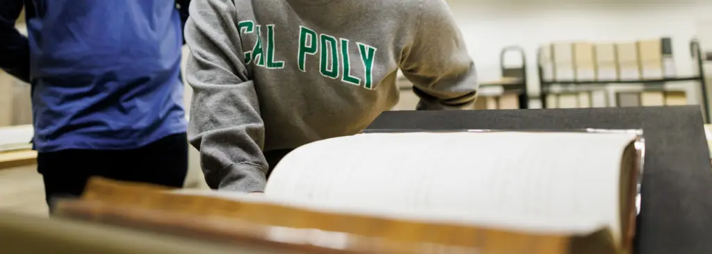 In the foreground, a 19th century ledger is on a book cradle. There are two people standing behind, looking at the ledger, just their torsos are visible, one is wearing a Cal Poly sweatshirt.